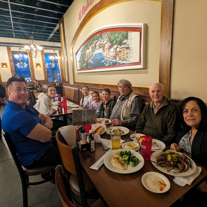 A family of seven enjoying dinner at Portofino's Greek and Italian Restaurant, with various Italian dishes on the table.