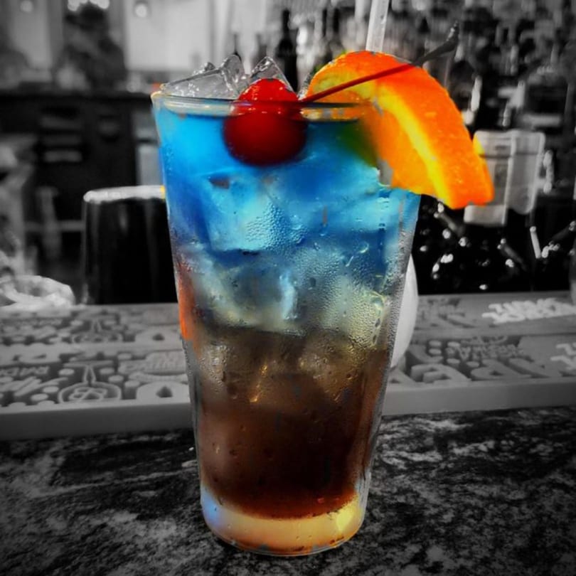 A vibrant blue and brown layered cocktail with ice, garnished with a cherry and orange slice.