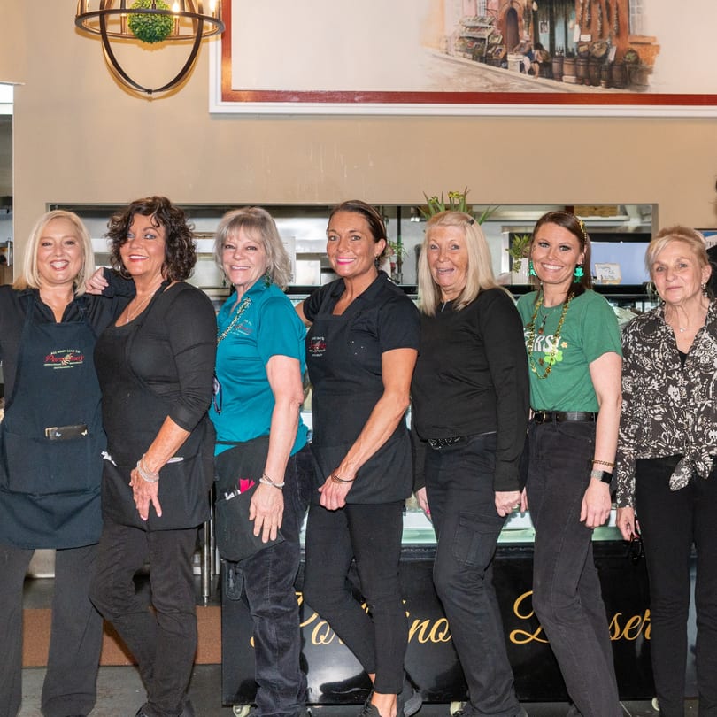 Six smiling women, Portofino's staff, stand together in front of a dessert display case.