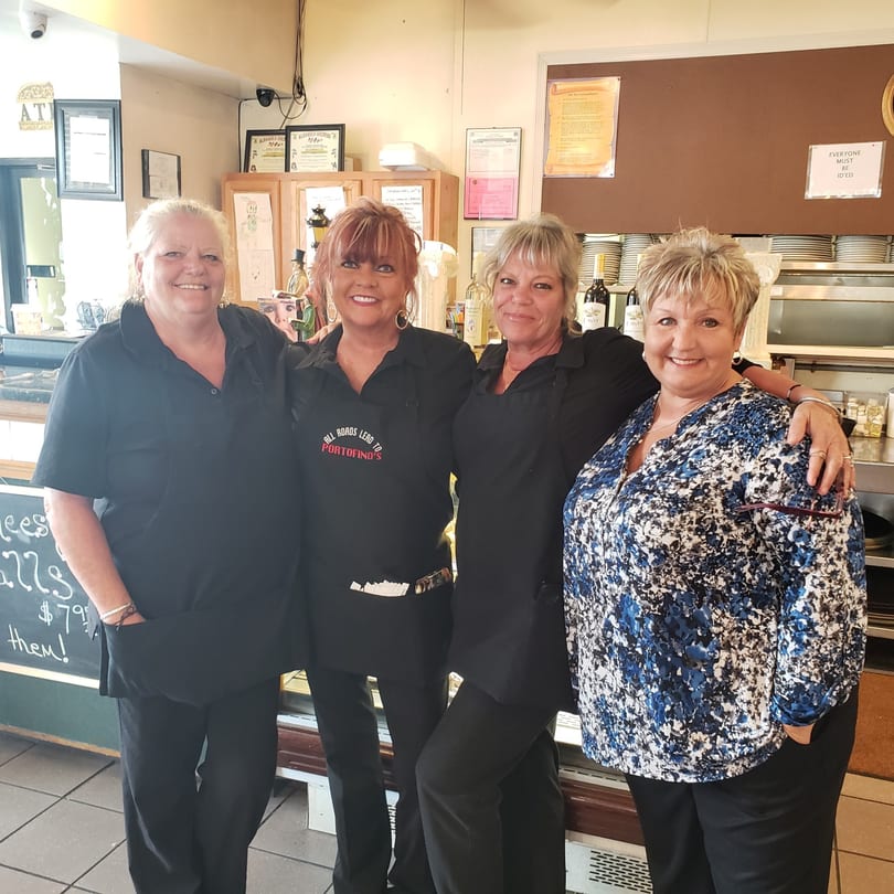 Four smiling women, three in black Portofino's aprons, stand together in a restaurant setting.