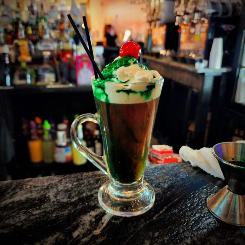 A green and white layered cocktail with a cherry and straws sits on a bar.