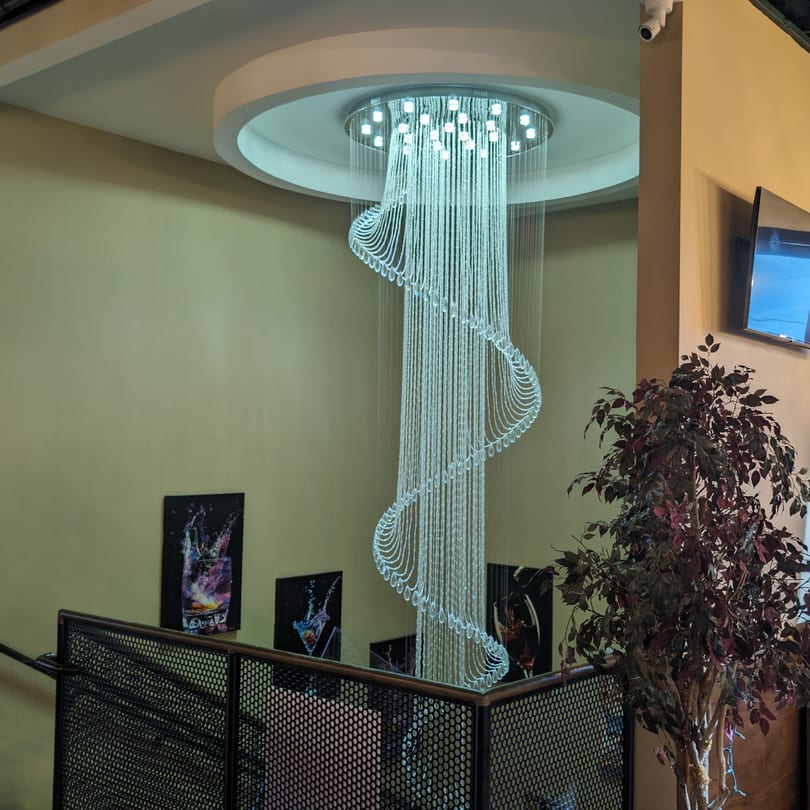A large, modern spiral chandelier hangs from the ceiling above a railing, with cocktail art on the wall.