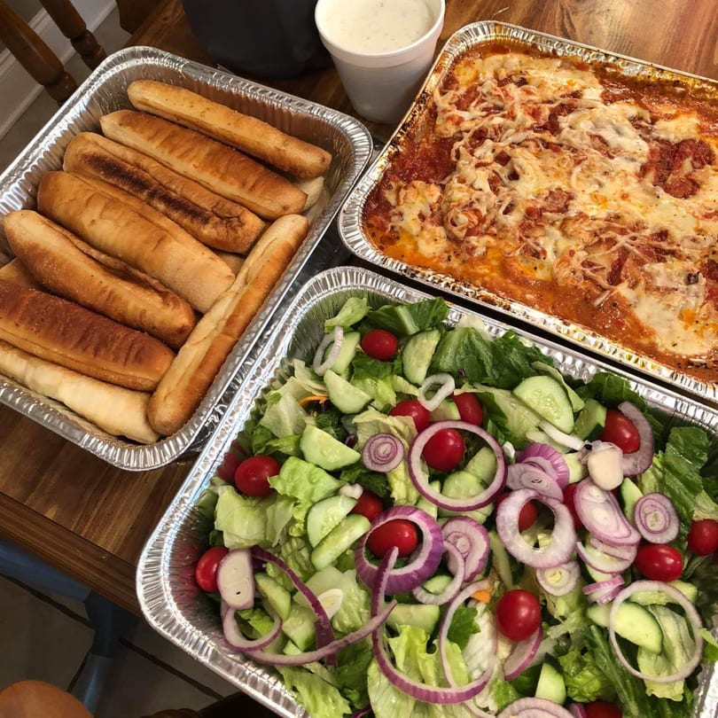 Three foil trays of Italian food: breadsticks, baked lasagna, and a fresh garden salad.