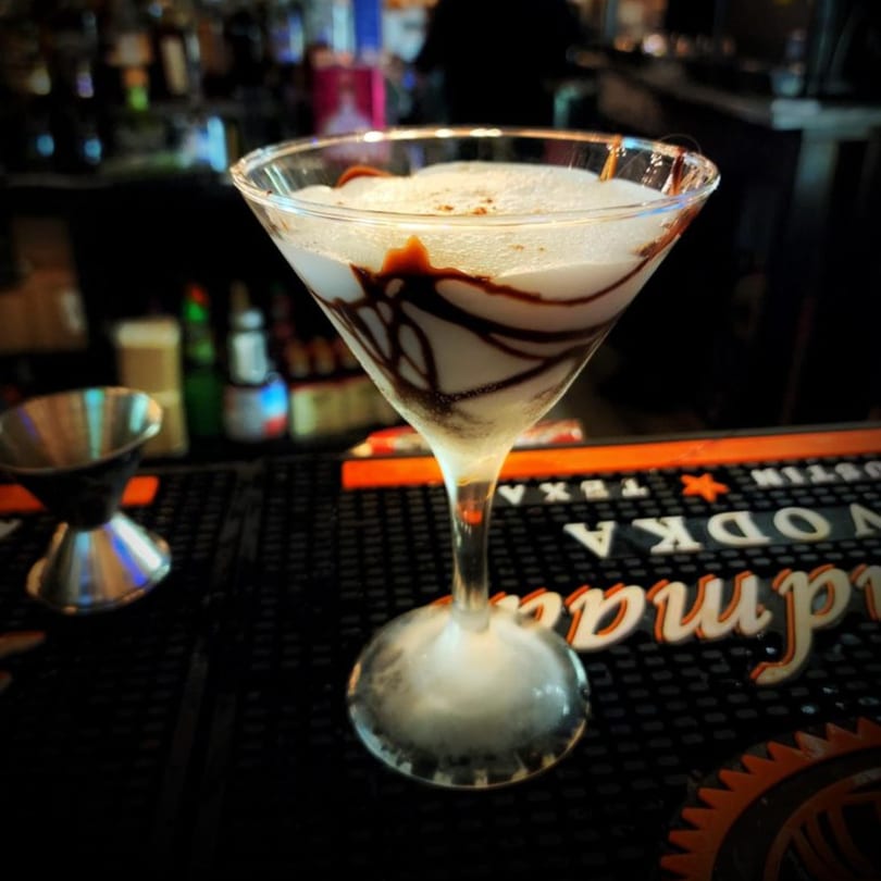 A white martini with chocolate drizzle and foam sits on a bar mat with bottles in the background.